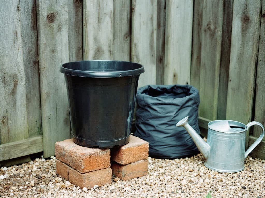 Empty black plastic pot with drainage holes and a fabric grow bag on a gravel patio, ready for growing potatoes.